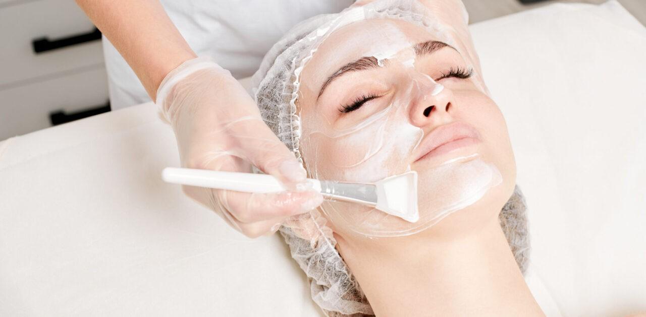 Beautician applies vitamins cream mask on woman face for rehydrate face skin, anti aging cosmetic procedure in beauty spa salon. Cosmetologist in gloves holds cosmetic brush for applying mask